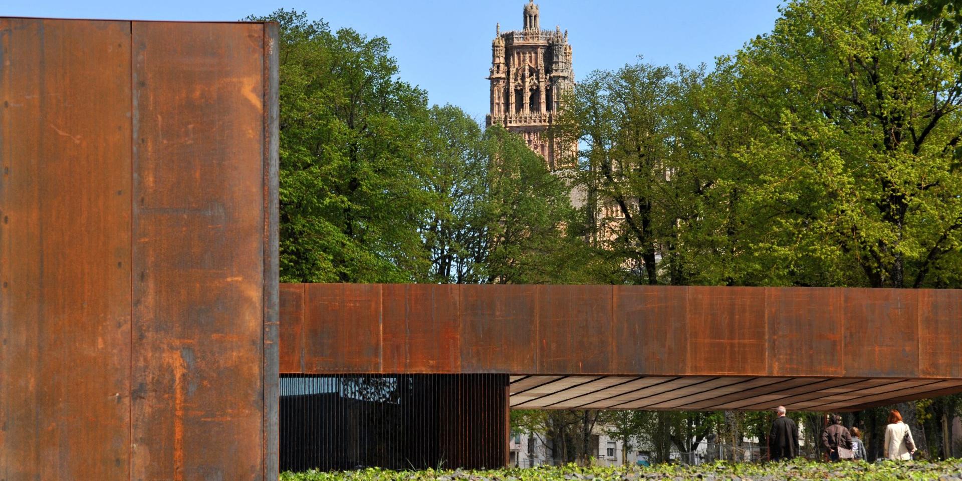 Hall d'entrée du musée Soulages en acier Corten vu de côté avec le clocher de la cathédrale de Rodez en arrière-plan entre les arbres