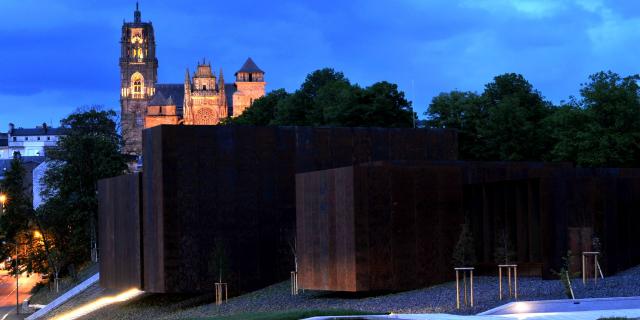 Vue de nuit du musée Soulages entouré d'arbres et de la cathédrale de Rodez en fond