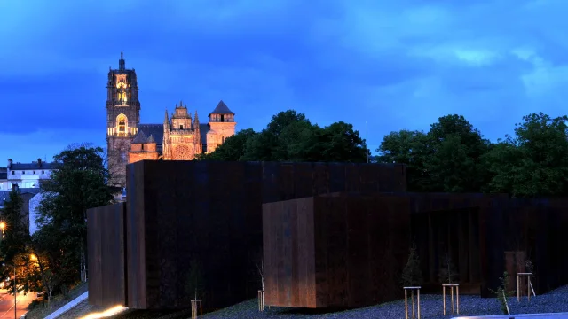Vue de nuit du musée Soulages entouré d'arbres et de la cathédrale de Rodez en fond
