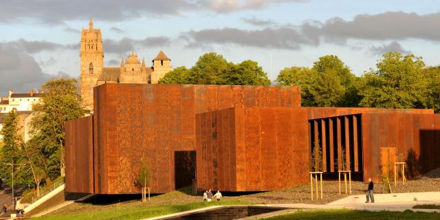 Vue du musée Soulages en acier Corten, passants en dessous et cathédrale en arrière-plan