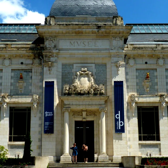 Façade principale du musée des Beaux-Arts Denys-Puech à Rodez, avec ses chiens jaunes sculptés et médaillons, devant un parking