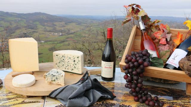 Plateau de fromages avec du Laguiole, Roquefort, Bleu des Causses, à côté d'une bouteille de Vin de Marcillac, et d'une caisse en bois avec deux autres bouteilles de vin et une grappe de raisins, face à une vue sur le vallon de Marcillac