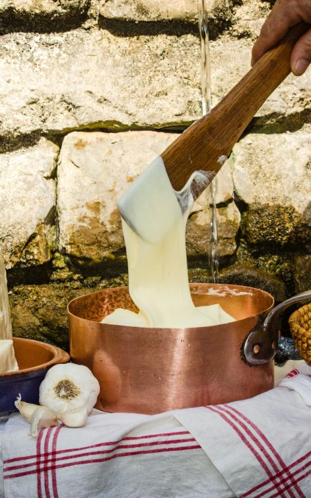 Devant un mur de pierres, sur des torchons, un plat en terre cuite avec deux morceaux de tomme, une casserole avec de l'aligot filant depuis une cuilllère en bois, et un plat en osier avec des pommes de terre, de l'ail