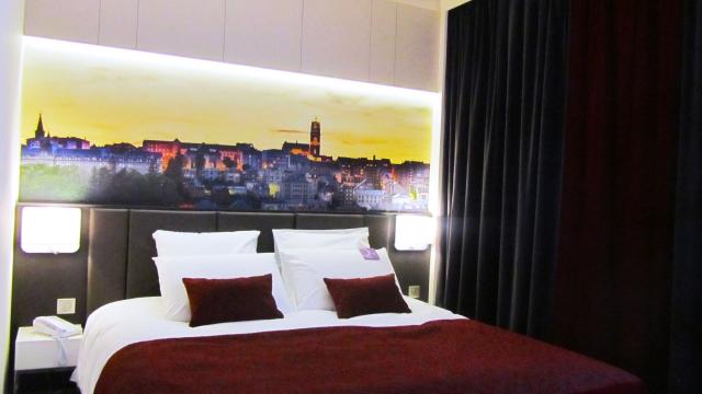 Lit double de l'Hôtel Mercure avec coussins blancs et par-dessus bordeaux avec une tête de lit en cuir noir, au-dessous un tableau panoramique de la ville de Rodez et sa cathédrale au coucher du soleil