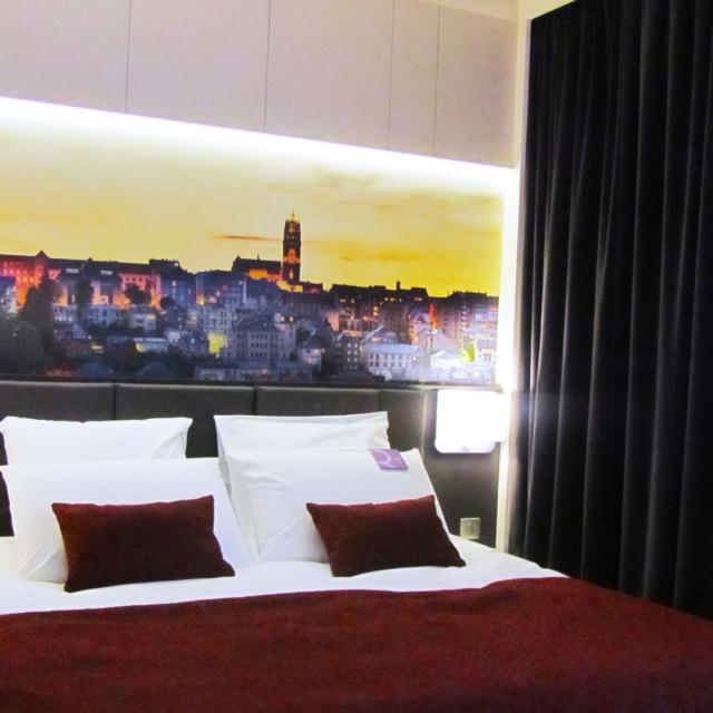 Lit double de l'Hôtel Mercure avec coussins blancs et par-dessus bordeaux avec une tête de lit en cuir noir, au-dessous un tableau panoramique de la ville de Rodez et sa cathédrale au coucher du soleil