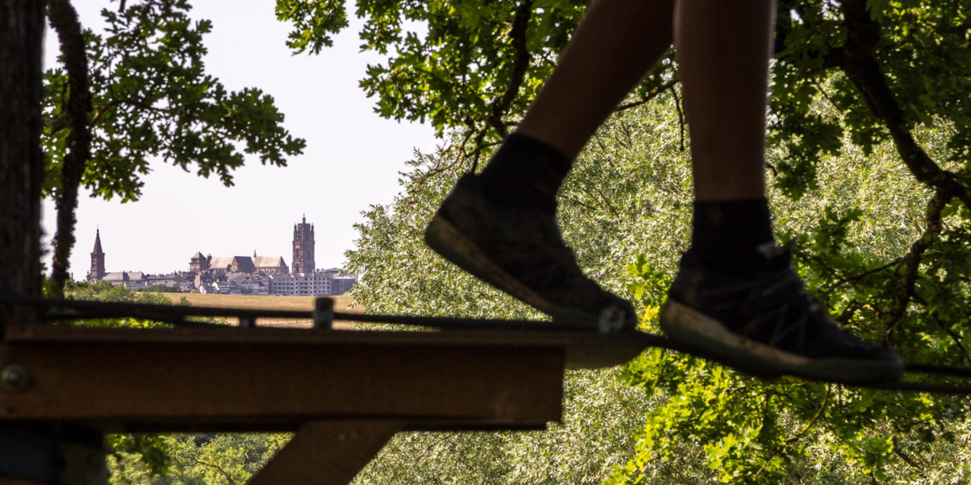 Paire de jambes en équilibre sur une corde dans les arbres lors d'un accrobranche, descendant d'un palier fixe en bois, avec une vue lointaine sur la cathédrale de Rodez