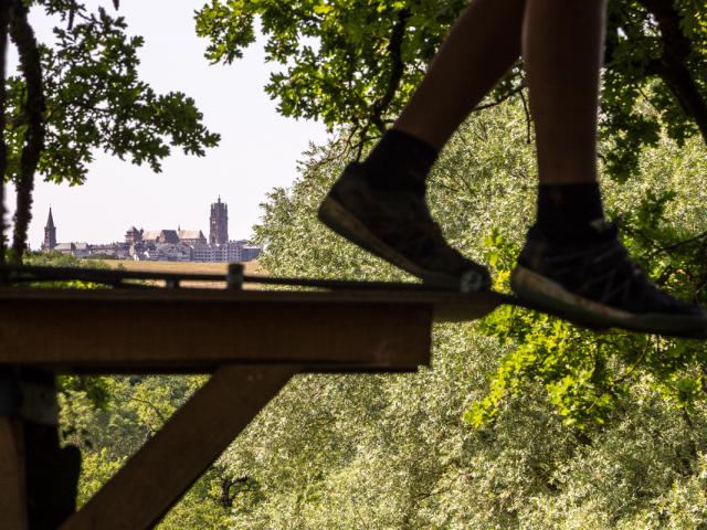 Paire de jambes en équilibre sur une corde dans les arbres lors d'un accrobranche, descendant d'un palier fixe en bois, avec une vue lointaine sur la cathédrale de Rodez