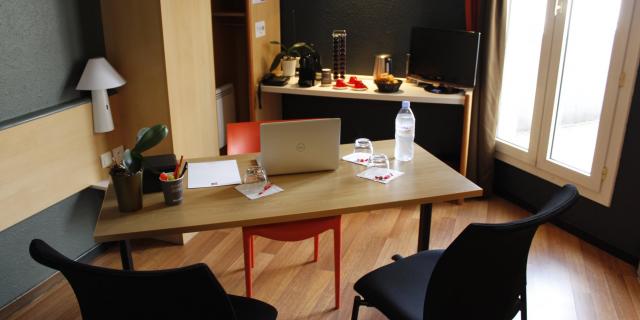 Bureau de l'hôtel Ibis avec table, chaises, ordinateur portable, bouteille d'eau et verres, cafetière et dosettes