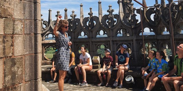 Groupe assis sur des rebords en pierre au sommet de la cathédrale, face à la guide-conférencière et dos aux détails sculptés du sommet