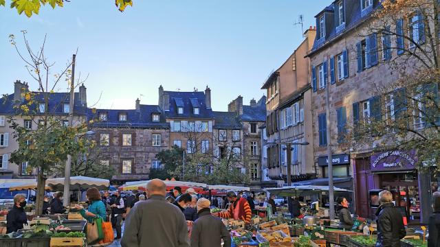 Personnes avec caddie marchant sur des feuilles mortes entre les étals de légumes, sur la place du marché devant les maisons typiques de Rodez