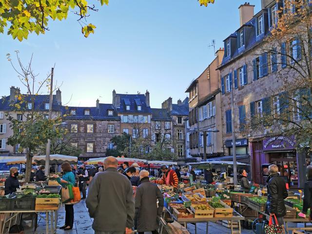 Personnes avec caddie marchant sur des feuilles mortes entre les étals de légumes, sur la place du marché devant les maisons typiques de Rodez