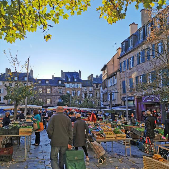 Personnes avec caddie marchant sur des feuilles mortes entre les étals de légumes, sur la place du marché devant les maisons typiques de Rodez