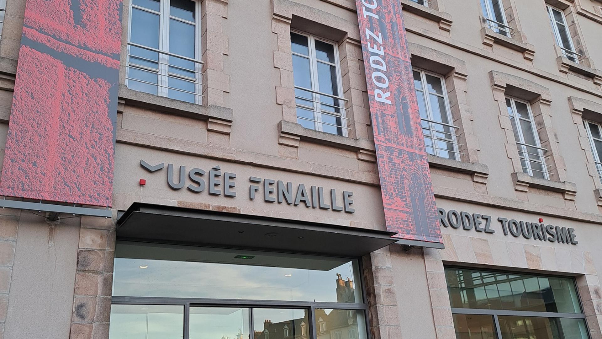 Devanture vitrée surmontée des enseignes Rodez Tourisme et musée Fenaille, agrémentées de bandeaux verticaux courant le long du bâtiment, avec le nom de l'enseigne et une photo associée, couleur rouge
