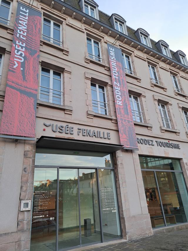 Devanture vitrée surmontée des enseignes Rodez Tourisme et musée Fenaille, agrémentées de bandeaux verticaux courant le long du bâtiment, avec le nom de l'enseigne et une photo associée, couleur rouge